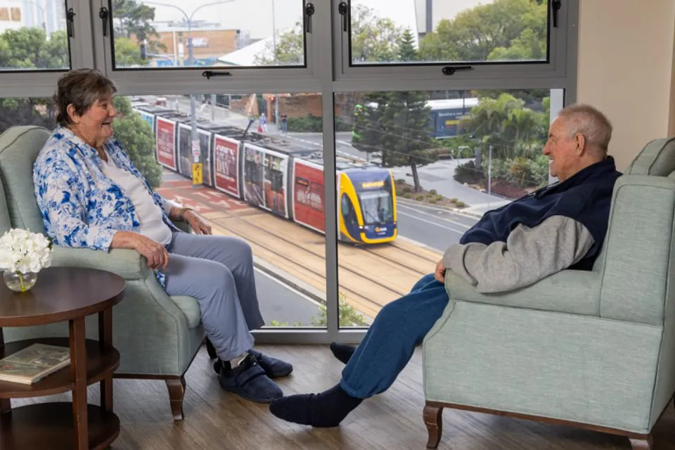 Two people seated in armchairs talking beside a large window
